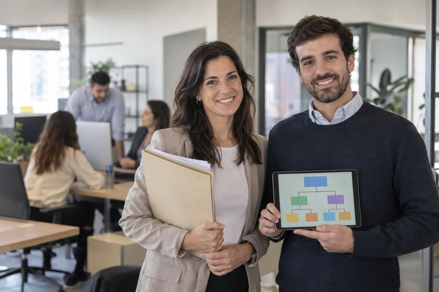 Responsable de recursos humanos con una carpeta de legajos y un analista con un organigrama en una tablet, en una oficina con empleados trabajando