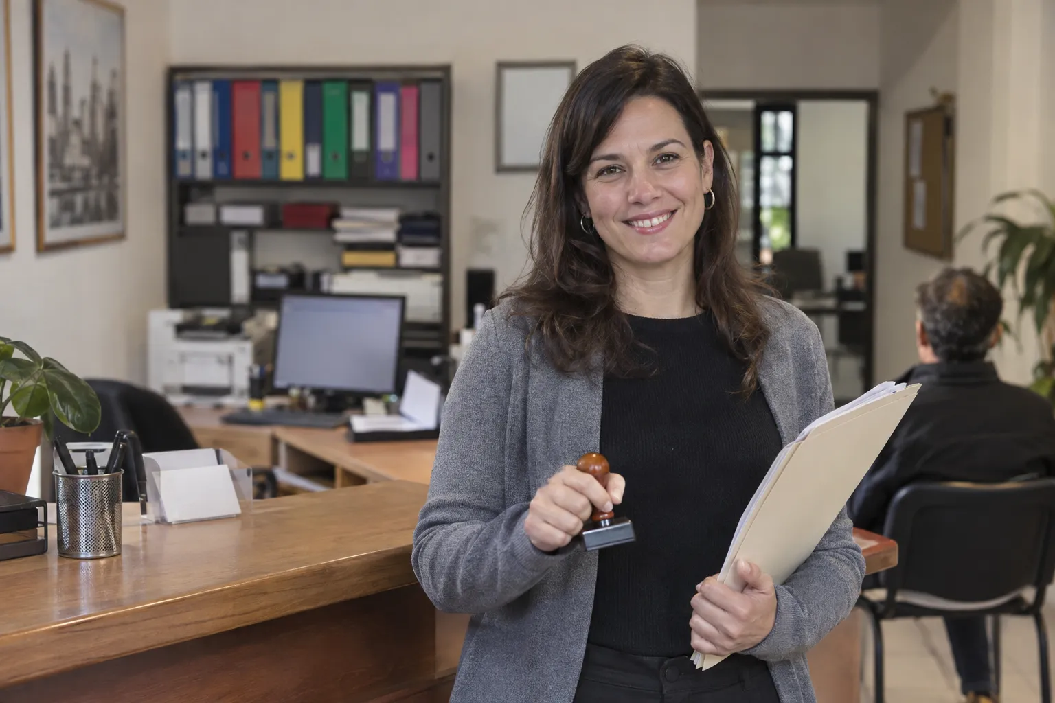 Gestora sonriente con un sello y una carpeta de trámites detrás del mostrador de una gestoría