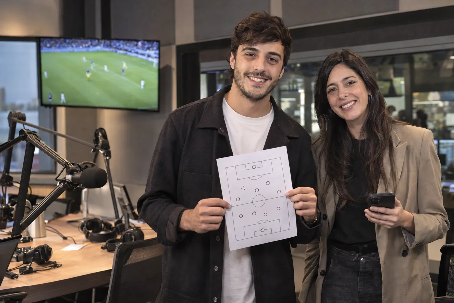 Dos periodistas deportivos sonrientes en un estudio de radio, él con la planilla de la formación de un equipo y ella con su celular, frente a micrófonos y un partido en pantalla